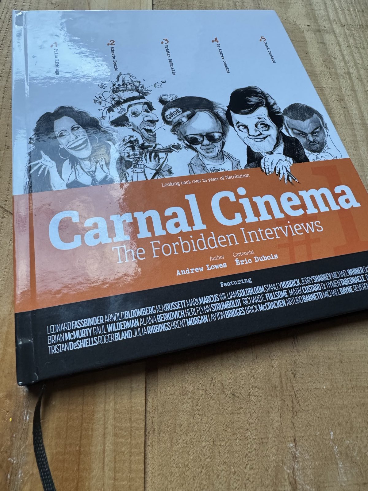 A photograph of the Carnal Cinema book on a wooden table. It's hard-backed with cartoons across the top, Carnal Cinema against Orange beneath that, and some tiny text beneath that. A dark grey ribbon emerges from the bottom.